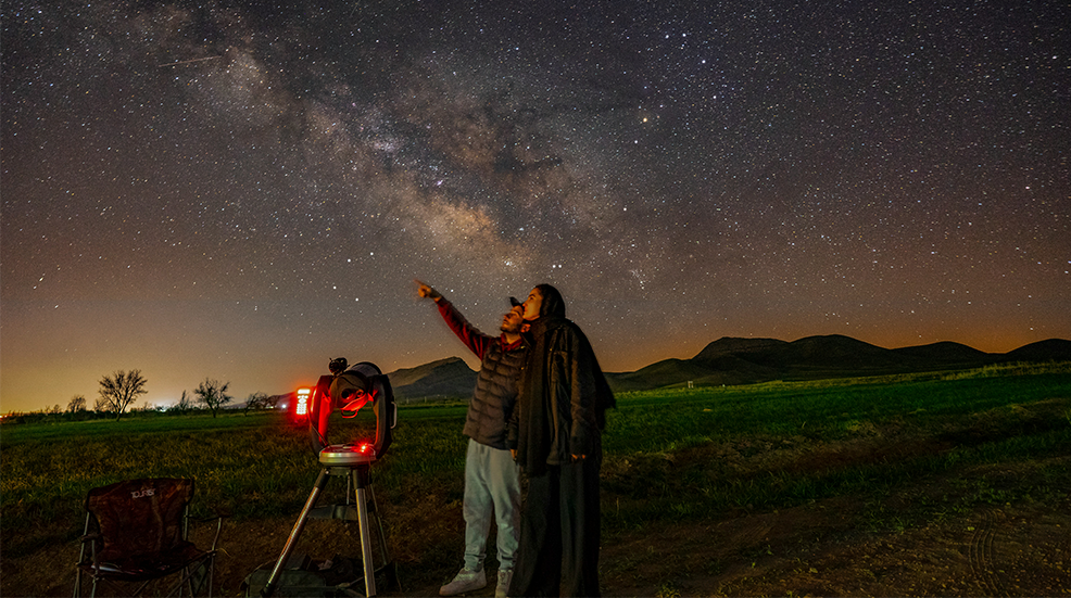 Two people with a telescope lookign up at a starry night sky